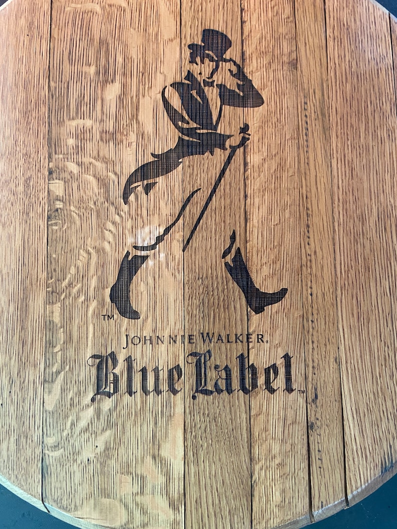 Pu&ograve; includere: Una botte di legno con un'immagine in bianco e nero di un uomo con un cappello a cilindro e un bastone. Il testo "Johnnie Walker Blue Label" &egrave; stampato sotto l'immagine.