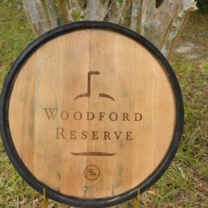 May include: Wooden barrel head with the words "Woodford Reserve" engraved in the center. The barrel head is circular with a dark rim and a light brown wood grain. A logo is above the text and a smaller logo is below.