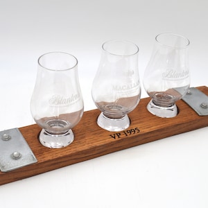 May include: A wooden tray with three small glasses for tasting whiskey. The tray is made from a reclaimed whiskey barrel and has metal accents. The glasses are engraved with the names of different whiskey brands: "Macallan", "Blanton's", and "Blanton's". The tray is engraved with the text "VP-1995".