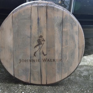 May include: A wooden barrel head with the Johnnie Walker logo and a walking man figure. The barrel head is brown and has a weathered look.