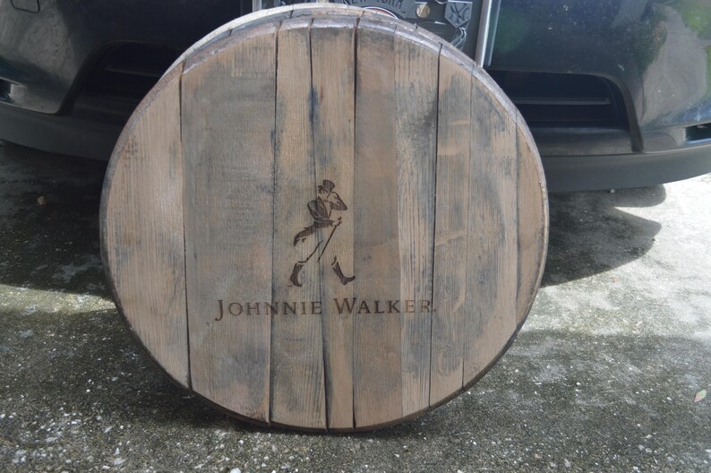 May include: A wooden barrel head with the Johnnie Walker logo and a walking man figure. The barrel head is brown and has a weathered look.