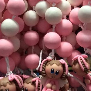 Puede incluir: Una cadena de cuentas rosadas y blancas con pequeñas figuras de ángeles adjuntas. Los ángeles llevan vestidos rosas y tienen cabello castaño y ojos azules.