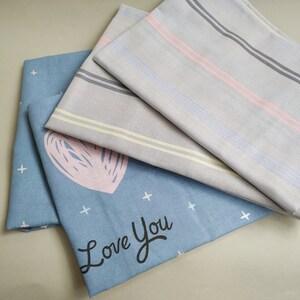 May include: A stack of three fabric squares. The top square is light gray with pink and blue stripes. The middle square is light gray with yellow stripes. The bottom square is light blue with white stars and the text "Love You" in black.