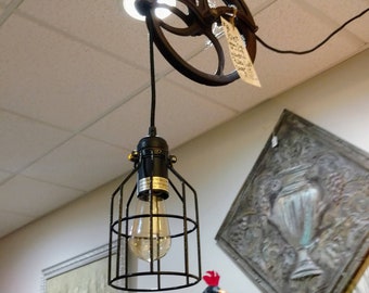 May include: A black caged pendant light hangs from a vintage metal pulley. The light is suspended from a ceiling with a white grid pattern. The pulley has a tag attached to it with text that is too blurry to read.