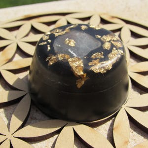May include: A black resin box with a clear lid and gold flakes embedded in the resin. The box is sitting on a wooden surface with a flower of life pattern.