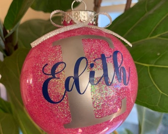 Può includere: Una palla di Natale rosa glitterata con il nome "Edith" in corsivo blu e una grande lettera "E" argentata. L'ornamento ha un cappuccio argentato e un fiocco di nastro argentato.