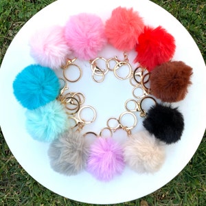 May include: A set of 10 colorful fluffy pom pom keychains with gold key rings. The pom poms are in various colors, including pink, red, blue, brown, black, and gray.