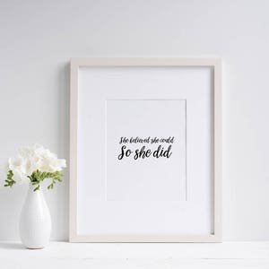 May include: A white framed wall art print featuring the black text "She believed she could So she did" centered on a white background, accompanied by a white vase containing a bouquet of white flowers.