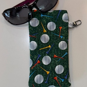 May include: A rectangular fabric pouch featuring a green background with a pattern of white golf balls and colorful golf tees, accompanied by a pair of dark-tinted sunglasses. The pouch includes a small silver metal clip attached to the side.