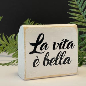 Könnte beinhalten: Ein weißes Holzschild mit schwarzer Aufschrift, die "La vita è bella" lautet.