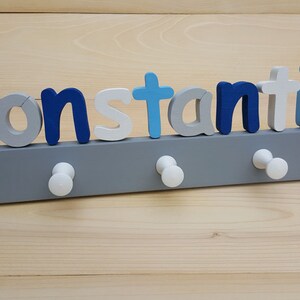 Peut inclure: Un porte-manteau en bois gris avec cinq crochets blancs. Le mot "Konstantin" est écrit en lettres de bois bleues, blanches et bleu clair au-dessus des crochets.