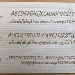 Op de afbeelding: Een close-up van een houten oppervlak met drie verschillende lettertypen weergegeven. De lettertypen zijn gelabeld "Schrift 1", "Schrift 2" en "Schrift 3". Elk lettertype toont het alfabet in hoofdletters en kleine letters, evenals de cijfers 1 tot en met 9 en 0.