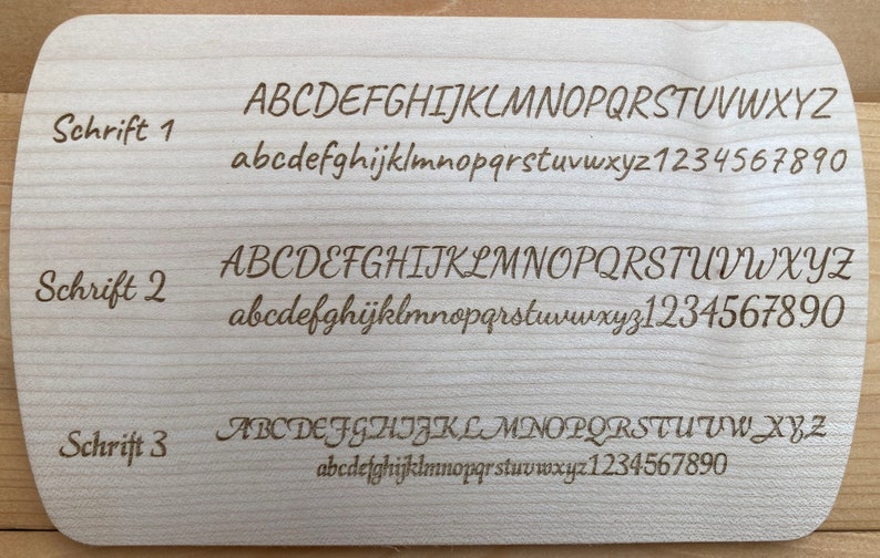 Op de afbeelding: Een close-up van een houten oppervlak met drie verschillende lettertypen weergegeven. De lettertypen zijn gelabeld "Schrift 1", "Schrift 2" en "Schrift 3". Elk lettertype toont het alfabet in hoofdletters en kleine letters, evenals de cijfers 1 tot en met 9 en 0.
