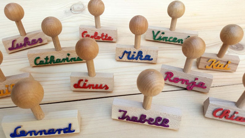 K&ouml;nnte beinhalten: Ein Set mit 10 Holzstempeln mit verschiedenen Namen, die in bunter Tinte geschrieben sind. Die Stempel sind aus Naturholz gefertigt und haben einen runden Griff. Die Namen auf den Stempeln sind: Lukas, Catharina, Linus, Mika, Vincent, Ronja, Kai, Isabelle, Chiara und Lennard.