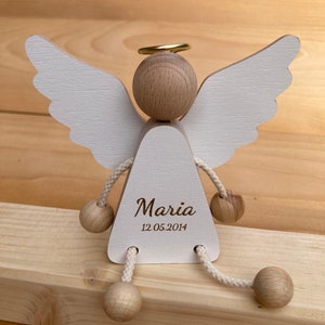 Op de afbeelding: Wit houten engelbeeldje met een halo, vleugels en een touwlichaam. De engel heeft een rond hoofd en een driehoekig lichaam met de naam "Maria" en de datum "12.05.2014" erop gegraveerd.