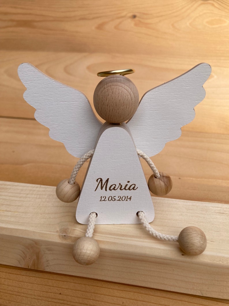 Op de afbeelding: Wit houten engelbeeldje met een halo, vleugels en een touwlichaam. De engel heeft een rond hoofd en een driehoekig lichaam met de naam "Maria" en de datum "12.05.2014" erop gegraveerd.
