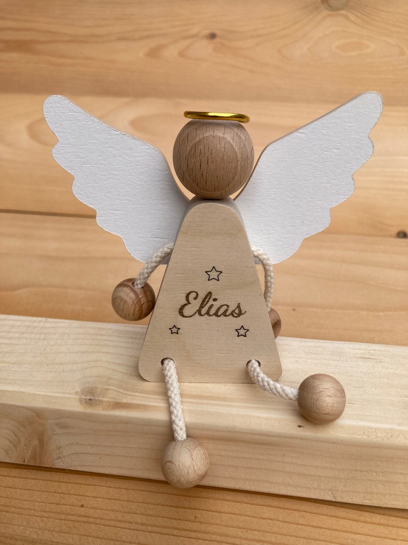 Op de afbeelding: Een houten engelbeeldje met witte vleugels, een gouden halo en de naam "Elias" gegraveerd op de voorkant. De engel heeft een driehoekig lichaam en vier houten ballen die aan touwtjes zijn bevestigd voor de benen.