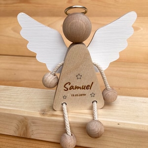Op de afbeelding: Houten engelbeeldje met witte vleugels, een halo en een touwtje dat aan zijn benen is bevestigd. De engel houdt een houten bal in elke hand. Het beeldje is gepersonaliseerd met de naam "Samuel" en de datum "12.05.2019".