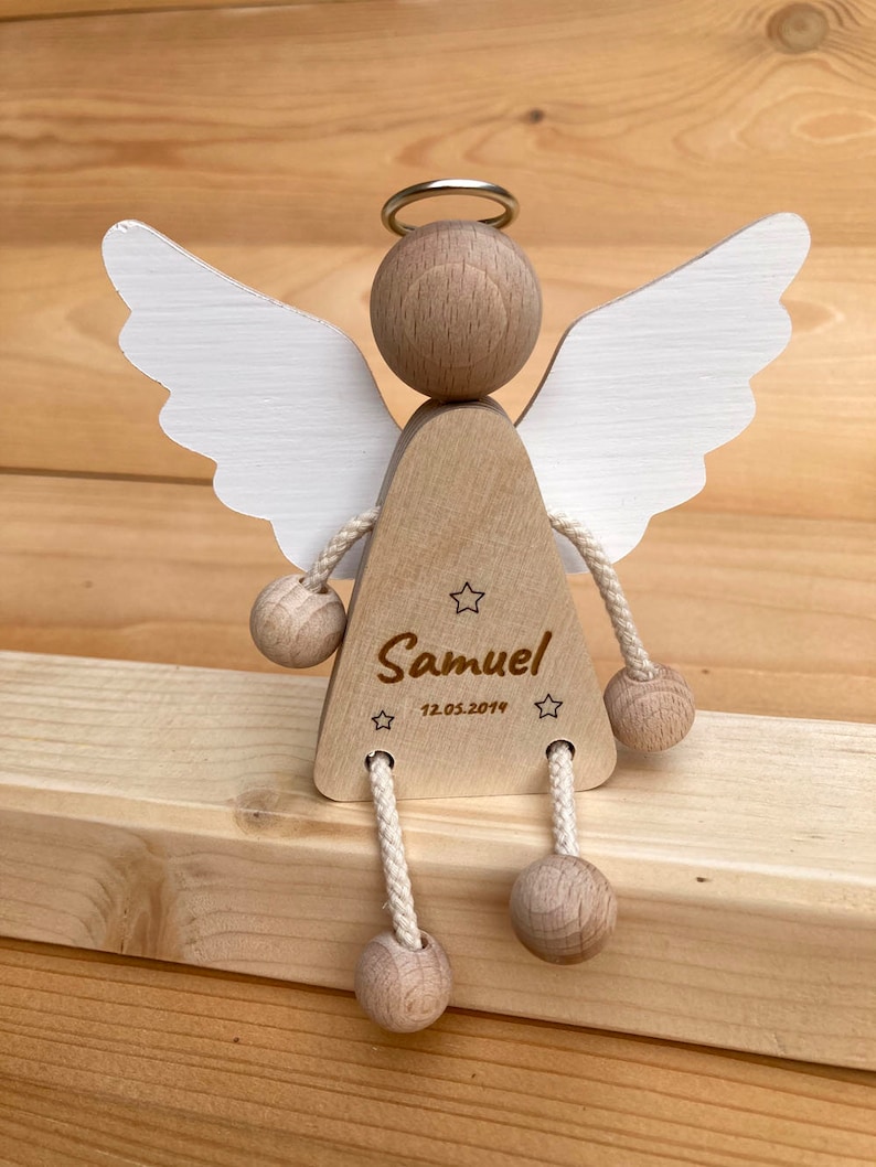 Op de afbeelding: Houten engelbeeldje met witte vleugels, een halo en een touwtje dat aan zijn benen is bevestigd. De engel houdt een houten bal in elke hand. Het beeldje is gepersonaliseerd met de naam "Samuel" en de datum "12.05.2019".