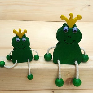 Könnte beinhalten: Zwei Holz-Froschfiguren mit gelben Kronen und blauen Augen. Der größere Frosch sitzt mit ausgestreckten Beinen, während der kleinere Frosch mit gebeugten Beinen sitzt. Beide Frösche sind grün bemalt.