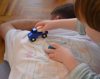 May include: A child's hands play with a blue toy cement mixer truck, featuring a white and orange cement mixer. The toy is placed on a white t-shirt with a colourful map design. The shirt is worn by a person lying down.