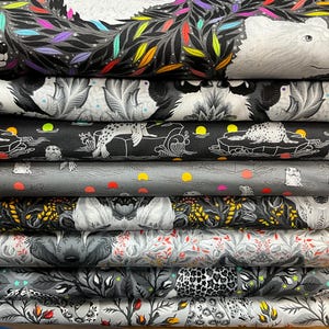 May include: A pile of patterned fabric rolls. The designs include a white polar bear with colourful leaves, seals, floral patterns, and animal illustrations. The colour palette is primarily black, white, and grey.