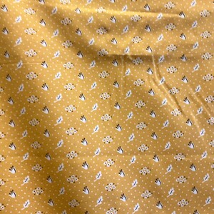 May include: Mustard yellow fabric with a repeating pattern of white dots, small floral clusters, and stylized leaf shapes in white and navy blue. The fabric has a slightly wrinkled texture.