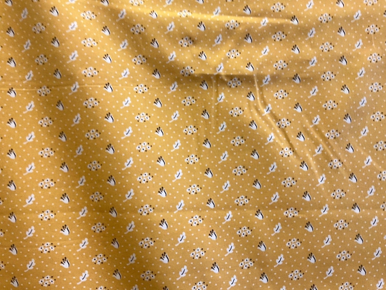 May include: Mustard yellow fabric with a repeating pattern of white dots, small floral clusters, and stylized leaf shapes in white and navy blue. The fabric has a slightly wrinkled texture.