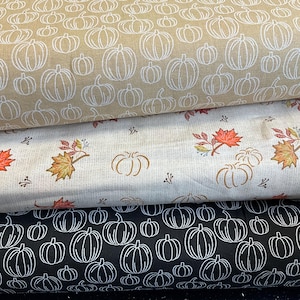 May include: Three rolls of fabric with autumn patterns. The top roll has a beige background with white pumpkin outlines. The middle roll has a cream background with pumpkins and autumn leaves. The bottom roll has a black background with white pumpkin outlines.