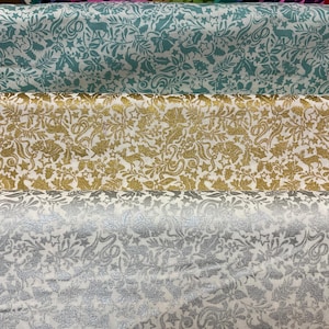 May include: Three rolls of white fabric with a repeating pattern of gold, silver, and teal glitter. The fabric has a festive, holiday theme.