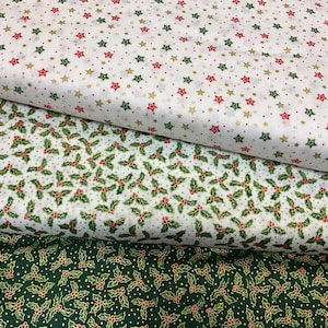 Może przedstawiać: Trzy rolki materiału o tematyce świątecznej. Górna rolka ma białe tło z czerwonymi, zielonymi i złotymi gwiazdami. Środkowa rolka ma białe tło z zielonymi liśćmi ostrokrzewu i czerwonymi jagodami. Dolna rolka jest ciemnozielona z ostrokrzewem.