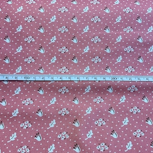 May include: Pink fabric with a repeating pattern of white flowers, leaves, and dots. A ruler is placed across the fabric, showing measurements in inches. The fabric has a vintage, floral design.