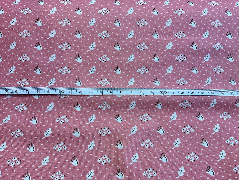 May include: Pink fabric with a repeating pattern of white flowers, leaves, and dots. A ruler is placed across the fabric, showing measurements in inches. The fabric has a vintage, floral design.