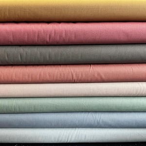 Puede incluir: Una pila de rollos de tela de varios colores. Los colores incluyen amarillo mostaza, rosa empolvado, gris, coral, rosa claro, verde salvia, azul claro y gris claro. La tela parece ser de algodón o lino tejido.