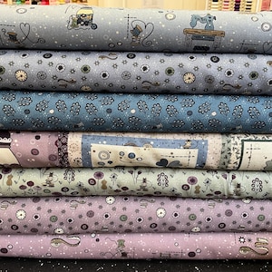 Może przedstawiać: Stos rolek materiału w różnych odcieniach niebieskiego, fioletowego i szarego. Tkaniny mają powtarzające się wzory maszyn do szycia, zegarów i motywów kwiatowych. Rolki są starannie ułożone, prezentując różne wzory i kolory.