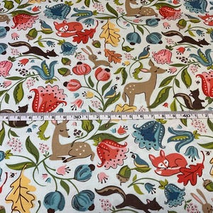 May include: White fabric with a whimsical forest pattern. Features deer, foxes, squirrels, rabbits, and birds among colorful flowers and foliage. A measuring tape is laid across the fabric, showing inches.
