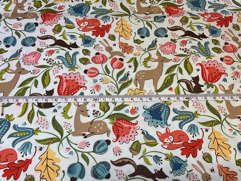 May include: White fabric with a whimsical forest pattern. Features deer, foxes, squirrels, rabbits, and birds among colorful flowers and foliage. A measuring tape is laid across the fabric, showing inches.