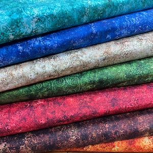 Puede incluir: Una pila de telas estampadas en varios colores, incluyendo turquesa, azul, beige, verde, rojo y marrón. Cada tela muestra un diseño floral o de remolinos detallado. Las telas están cuidadosamente dobladas, mostrando la variedad de colores y patrones.