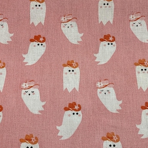 May include: Pink fabric with a repeating pattern of white ghosts wearing orange cowboy hats. The ghosts have black eyes and are smiling. The fabric has a textured appearance.