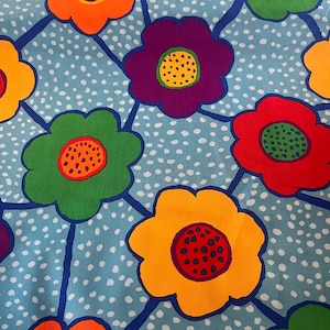 May include: A close-up of a fabric with a repeating floral pattern. The flowers are in shades of orange, yellow, purple, red, and green, with contrasting centers. The background is a light blue with white polka dots and dark blue lines.