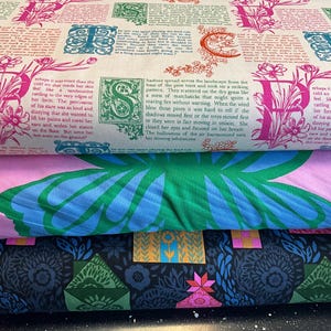 Può includere: Una pila di rotoli di tessuto fantasia. Il rotolo superiore presenta uno sfondo color crema con grandi lettere rosa e disegni floreali, insieme al testo. Il secondo rotolo ha uno sfondo rosa con un grande disegno di farfalla verde e blu. Il rotolo inferiore è blu scuro con motivi floreali e geometrici.