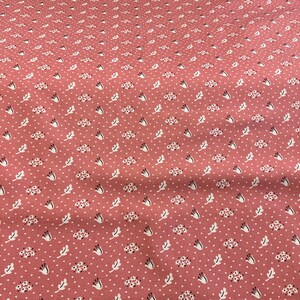 May include: A close-up of a pink fabric with a repeating pattern of white dots, small floral clusters, and stylized leaves. The fabric has a slightly textured appearance, suggesting a cotton or linen material.