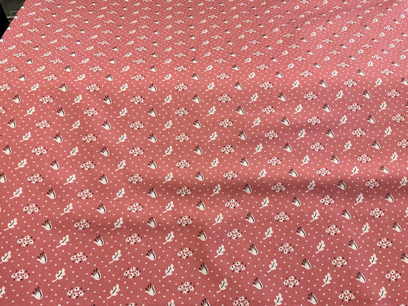 May include: A close-up of a pink fabric with a repeating pattern of white dots, small floral clusters, and stylized leaves. The fabric has a slightly textured appearance, suggesting a cotton or linen material.
