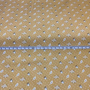 May include: Mustard-colored fabric with a repeating pattern of white dots, small floral clusters, and stylized white leaves. A blue and white measuring tape is laid across the fabric, showing measurements in inches.