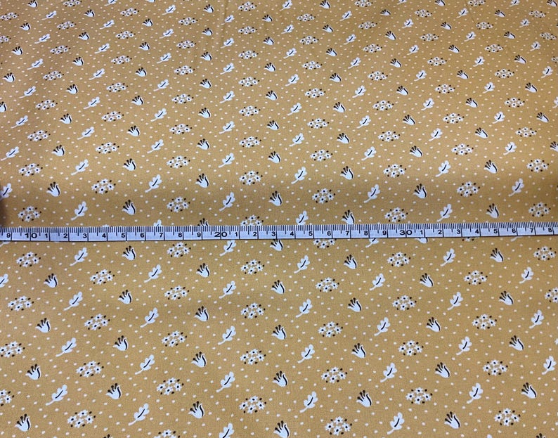 May include: Mustard-colored fabric with a repeating pattern of white dots, small floral clusters, and stylized white leaves. A blue and white measuring tape is laid across the fabric, showing measurements in inches.