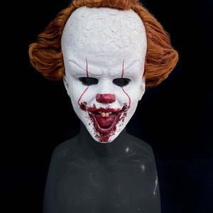 Könnte beinhalten: Eine weiße und rote Clownsmaske mit einem breiten, zahnigen Grinsen und Blut, das aus dem Mund tropft. Die Maske hat rote Linien, die auf das Gesicht gemalt sind, und eine rote Nase. Die Maske befindet sich auf einem schwarzen Mannequin-Kopf mit roten Haaren.