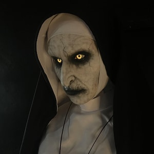 May include: A detailed mask of a nun with a pale complexion, cracked skin, and glowing yellow eyes. The nun's habit is white with a black veil. The image is set against a dark background, creating a spooky and eerie atmosphere.