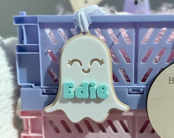 May include: A Halloween-themed display featuring a light blue and pink collapsible storage crate. The crate holds a ghost-shaped tag with the name "Edie" in blue letters. Ghost-shaped candles in pink and purple are in front of the crate.