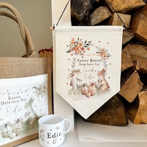 May include: A burlap Easter gift bag with a personalised bunny design and the text "Easter Delivery For Edie". A white ceramic mug with the name "Edie" and a floral design. A burlap banner with the text "Easter Bunny Stop here for Jacob & Edie" and a floral design.
