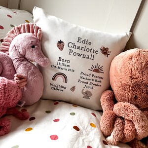 Könnte beinhalten: Ein weißes Kissen mit brauner Stickerei, auf dem "Edie Charlotte Pownall" steht, sowie eine Geburtsanzeige und Details. Das Kissen ist mit einer Erdbeere, einer Blume, einem Regenbogen, einer Sonne, einem Blatt und einem Elefanten verziert.